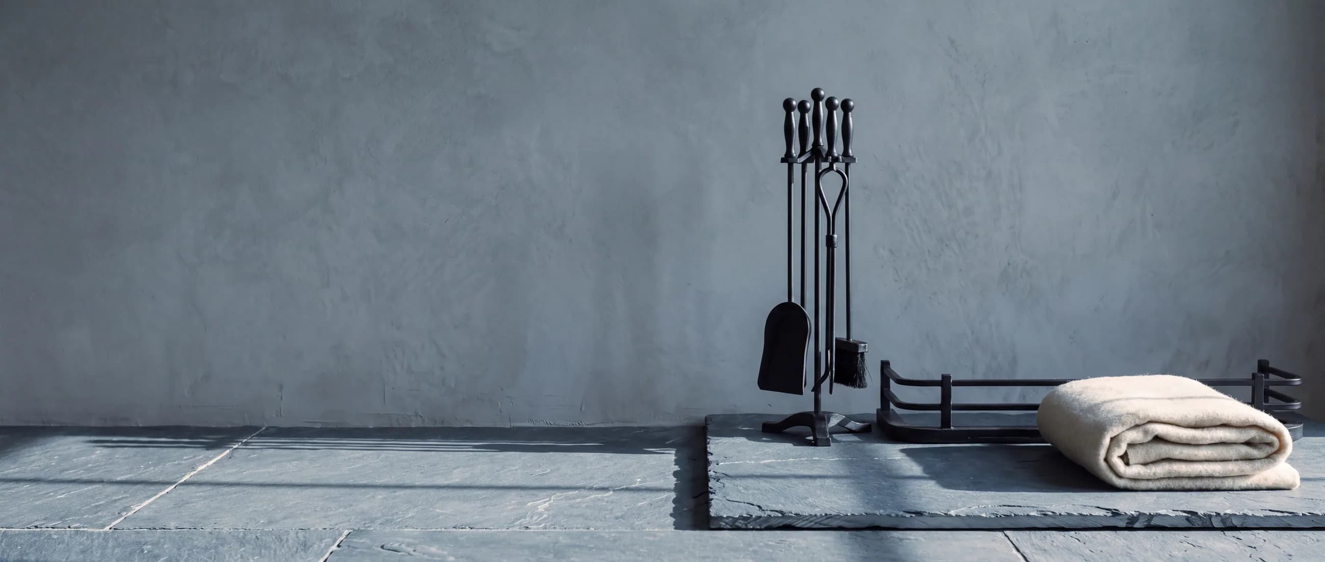 A four-piece blackened steel fire tool set, a low steel hearth fender and a folded undyed wool blanket on a riven slate hearth in cold winter light.