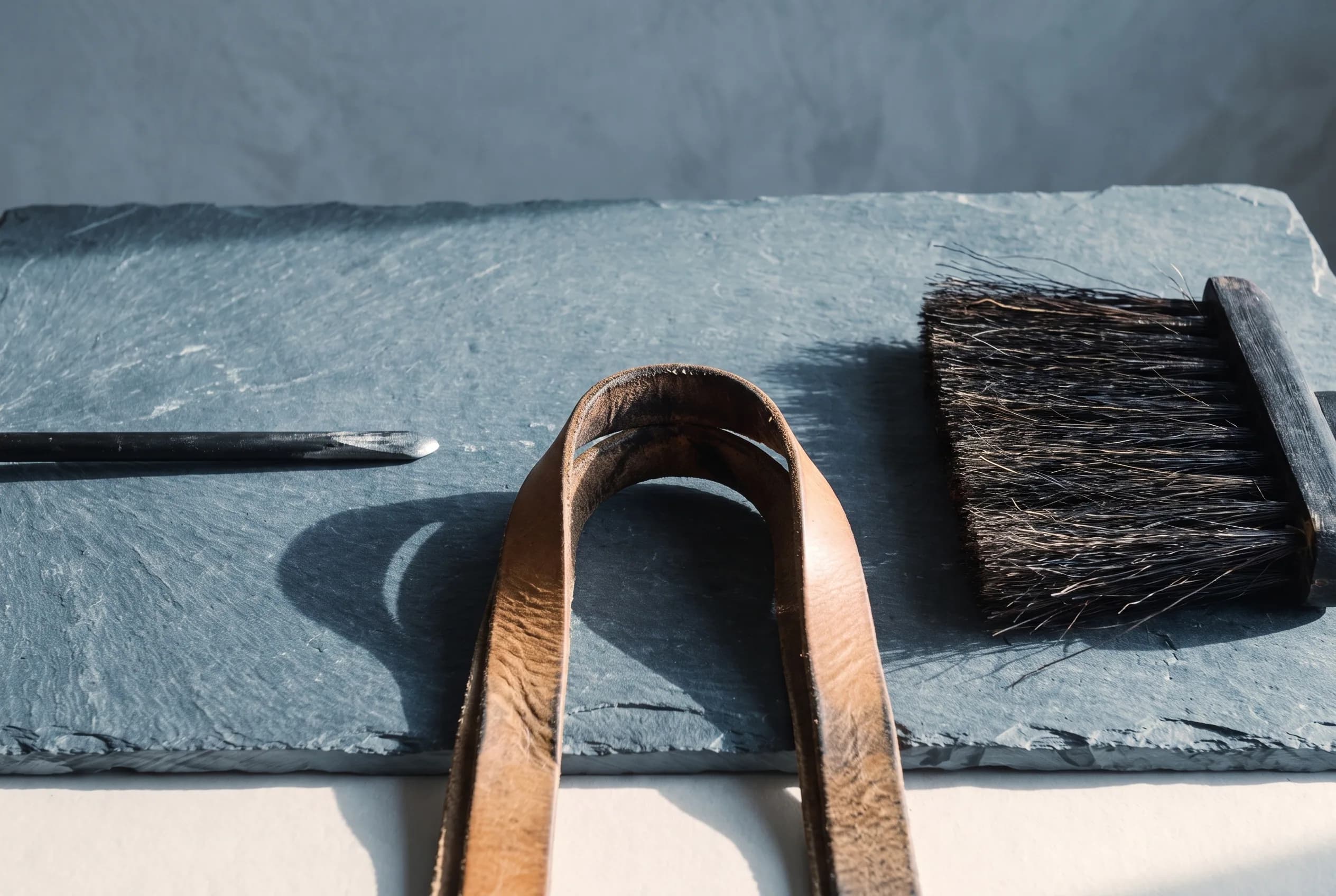 Three pieces photographed at their wear points: a poker whose black finish has worn to bright graphite at the tip, a log carrier handle whose leather has darkened and creased at the fold, and a horsehair broom head worn short along its leading edge.