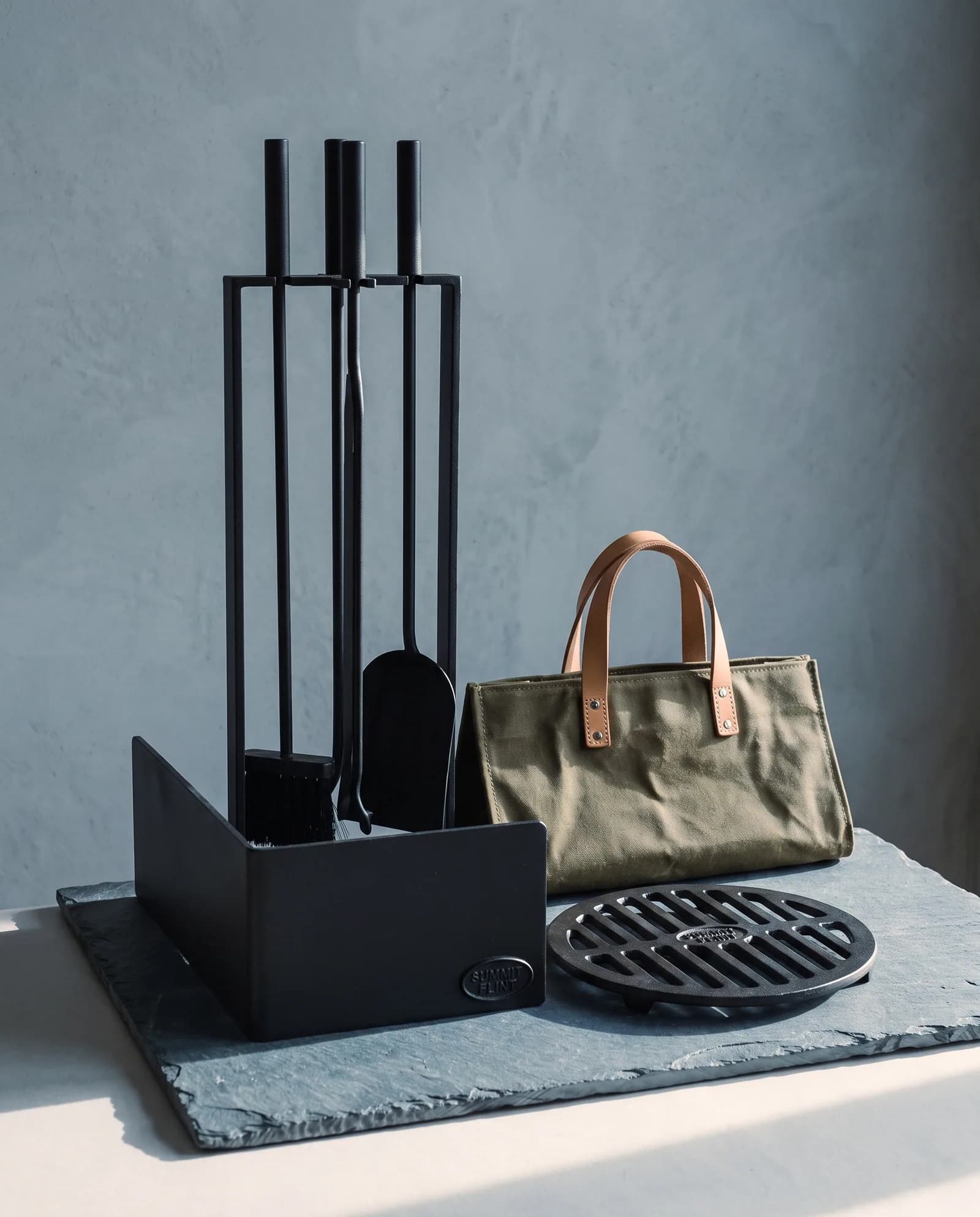 The Hearth set: a standing four-piece steel fire tool set, a low steel fender, a waxed canvas log carrier and a cast iron trivet, grouped on riven slate.